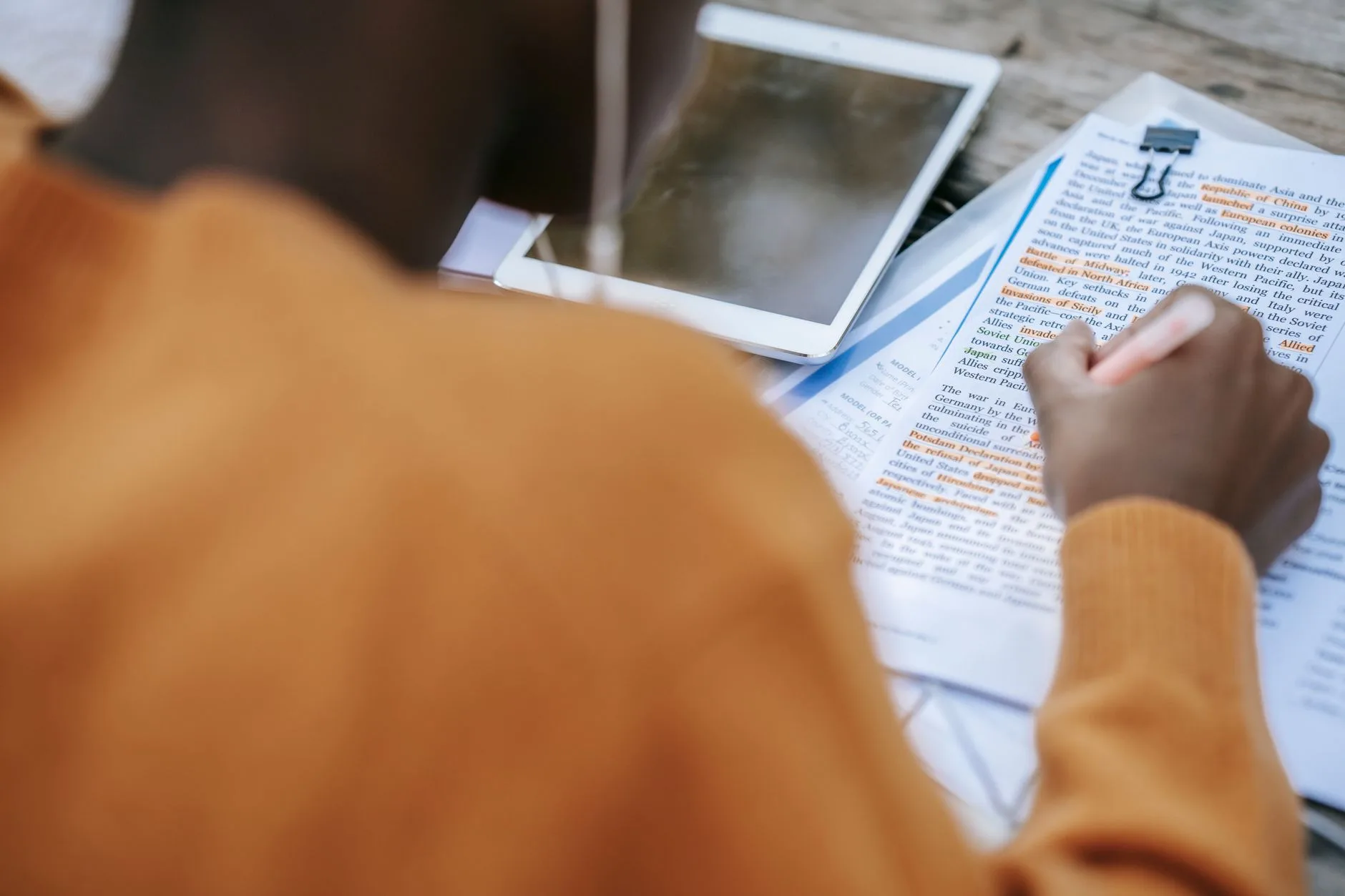 African American student annotating printed text at a desk with a tablet nearby, viewed from above